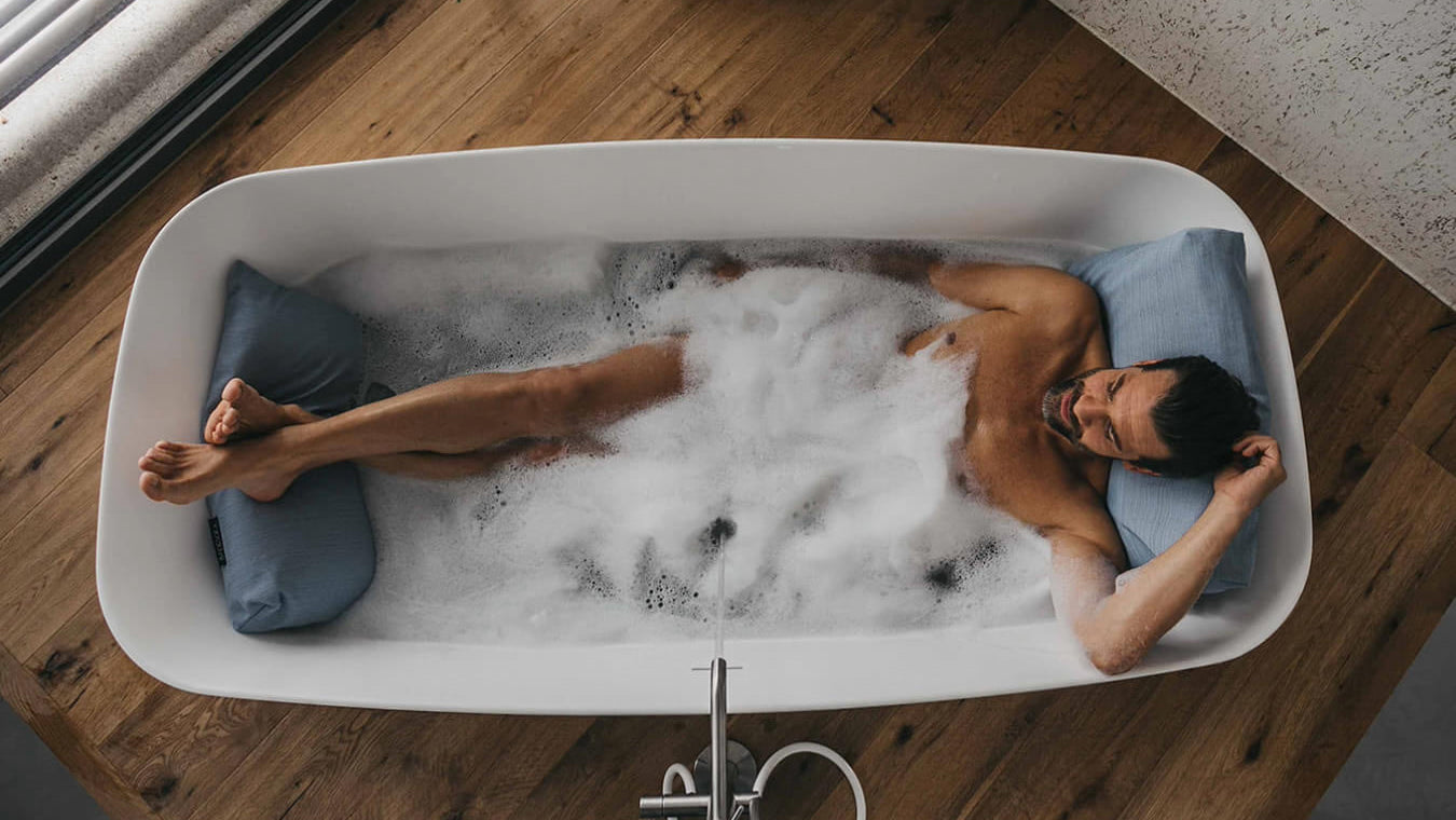 Mann badet mit Rückenkissen (Large, Sky oder Hellblau) und Fusskissen in luxuriöser freistehender Badewanne. Rücken, Kopf und Füsse ruhen entspannt auf Badesofas (Farbe Sky oder Hellblau). $ Freistehende Badewanne | Size Large Set | Sky