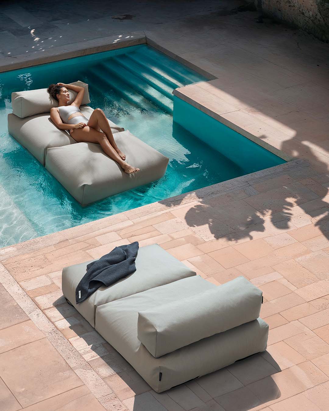 Eine Frau liegt entspannt auf einem Poolsofa double in Beige, das im Pool schwimmt. Am Rand des Pools, auf der Terrasse steht ein weiteres Poolsofa. $ Poolsofa Double | Floating Lounger | Beige