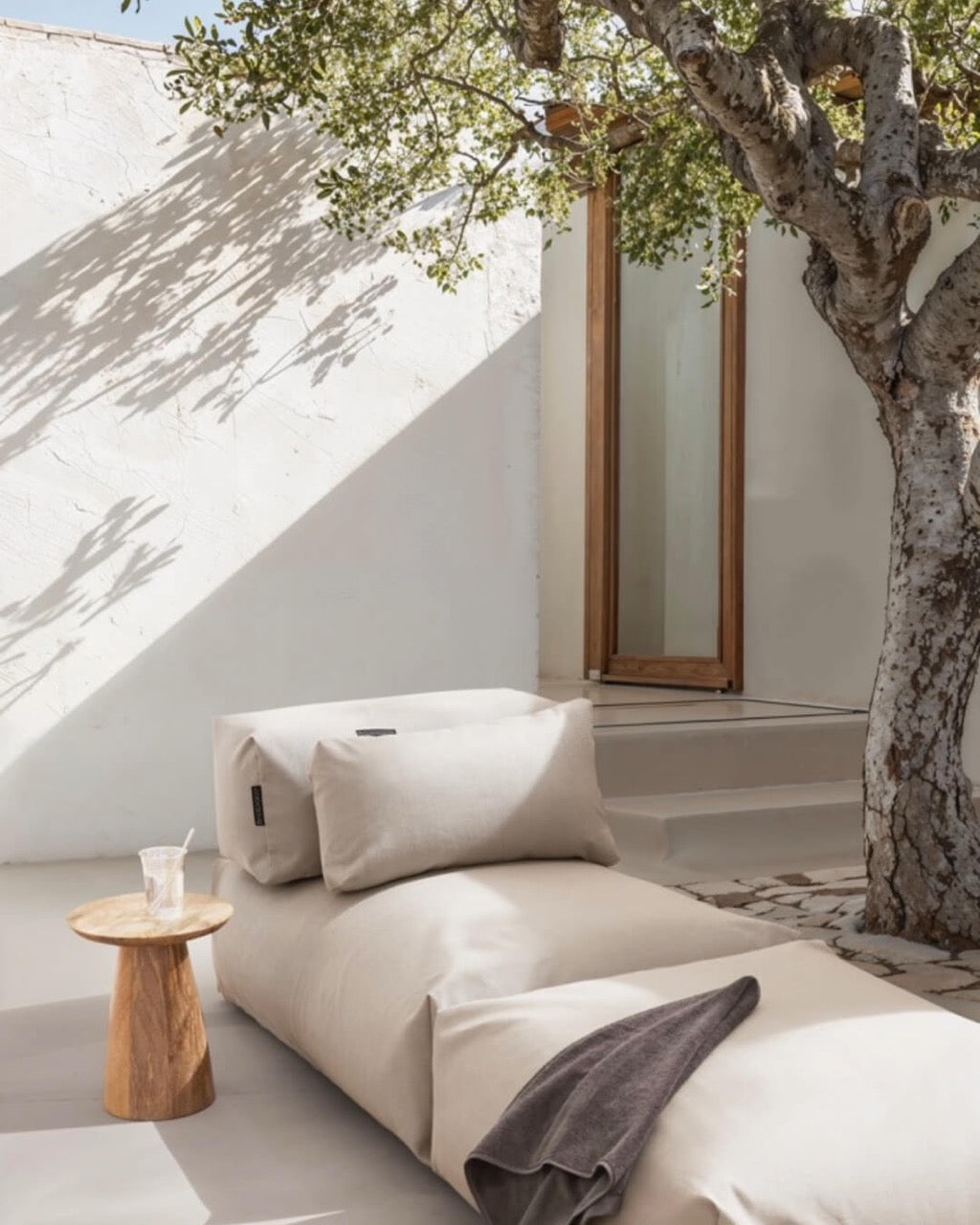 Unter einem Baum steht ein Poolsofa Singel in Beige, daneben ein kleiner Holztisch. Das Rückenkissen und Relaxkissen sind auf dem Poolsofa drapiert.