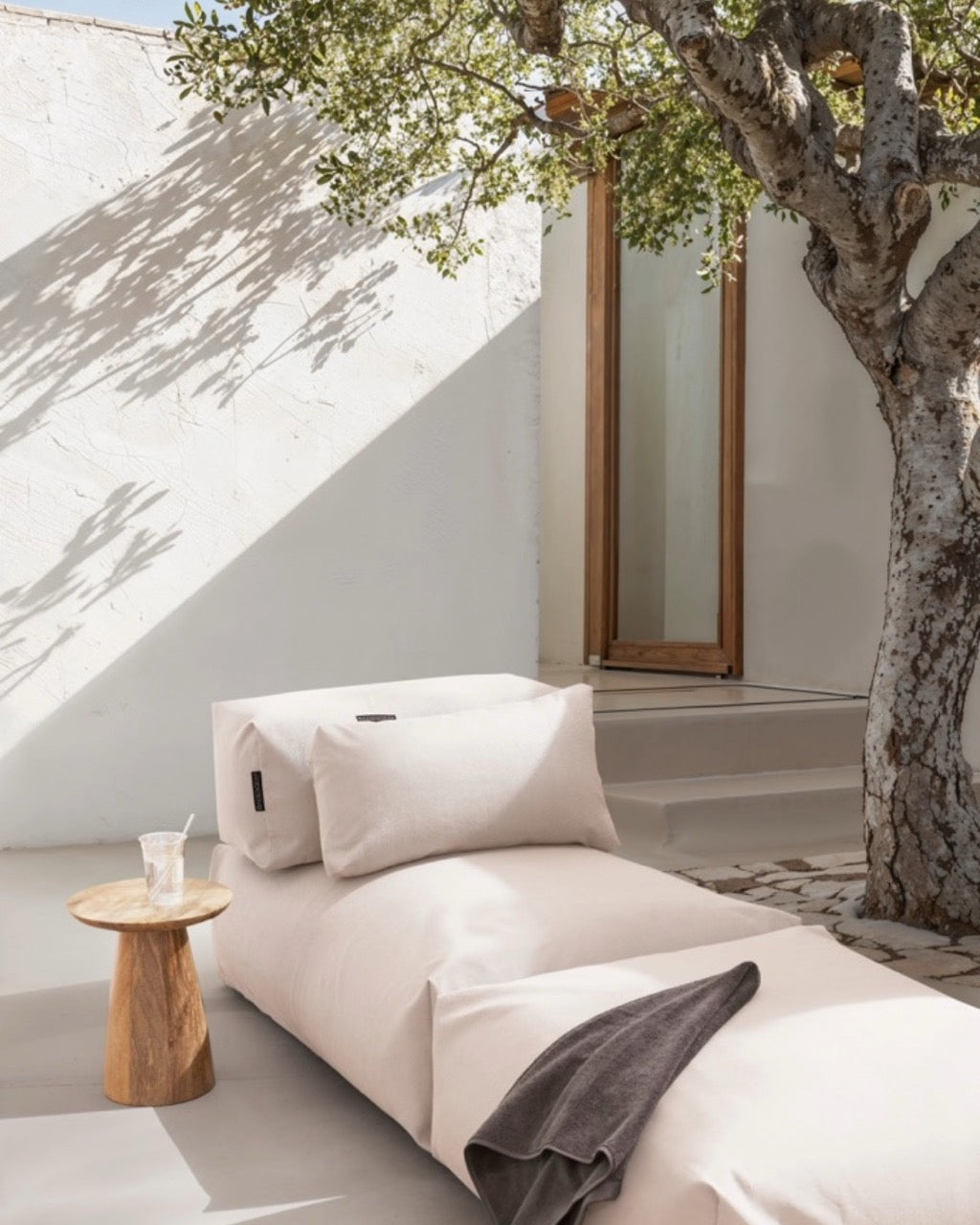 Unter einem Baum steht ein Poolsofa Singel in Perlmutt, daneben ein kleiner Holztisch. Das Rückenkissen und Relaxkissen sind auf dem Poolsofa drapiert. #farbe_perlmutt