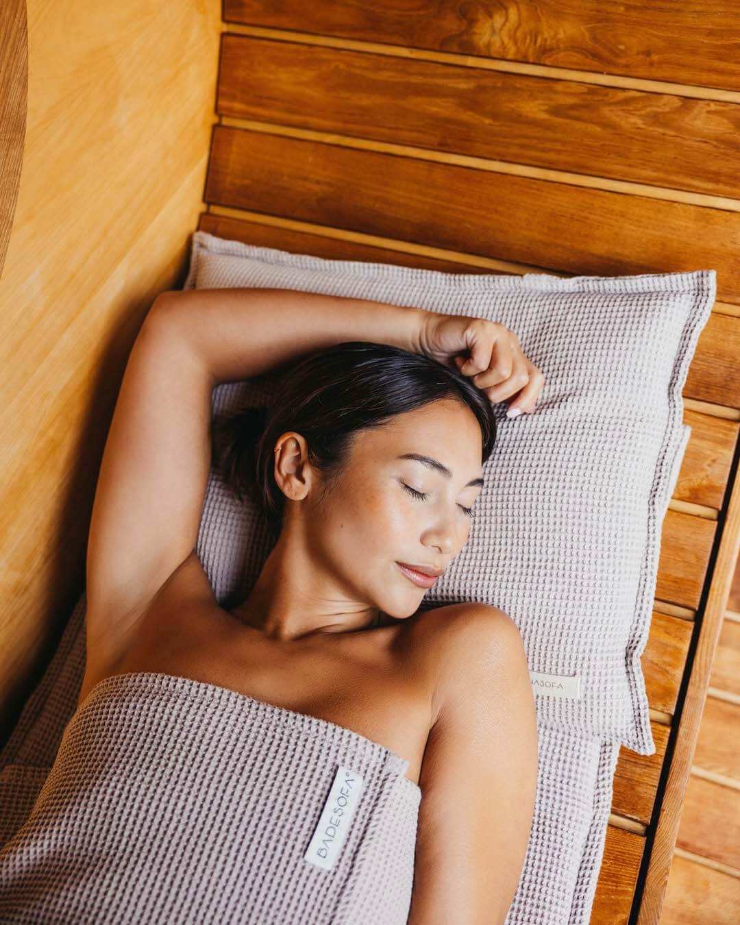 Frau liegt entspannt auf einem Saunasofa Kopfkissen und der gepolsterten Saunaauflage in Taupe.