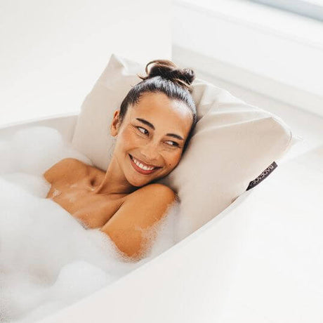 Eine Frau liegt lächelnd in einer freistehenden Badewanne, sie lehnt sich an ein Badesofa Rückenkissen Large in der Farbe Perlmutt.