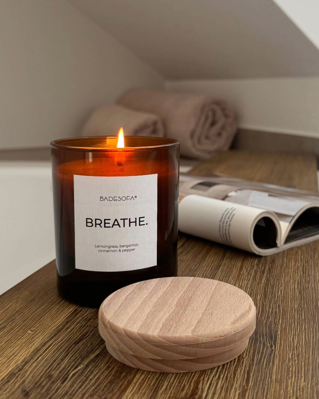 Brennende Duftkerze steht am Badewannenrand. Der Holzdeckel liegt daneben. 